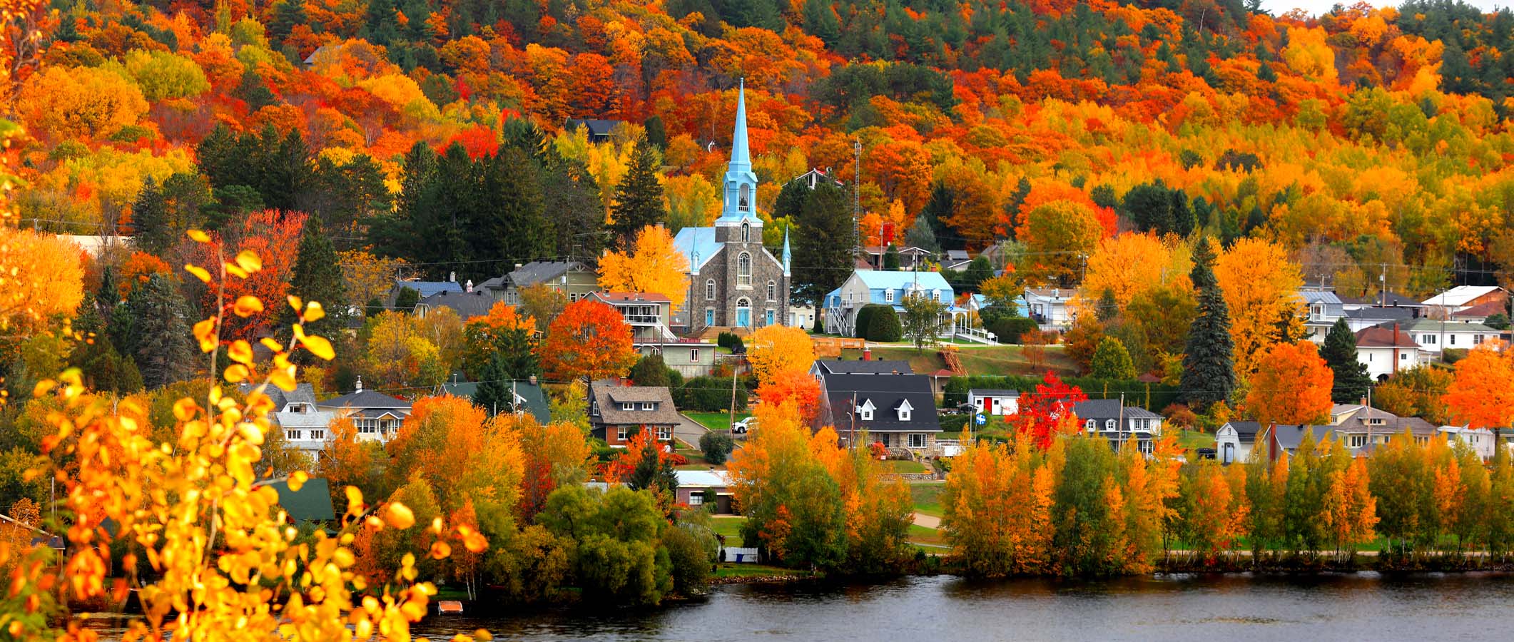 Vue frontale d'un paysage québécois automnal où, au premier plan, on peut aprecevoir une église au bord de l'eau et entourée d'arbres aux couleurs vives